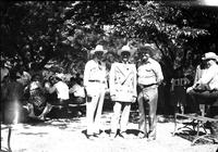 [Autry standing with two other men in front of people at picnic tables in park]