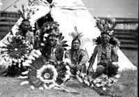 [Four Indian men in native dress sitting and kneeling in front of tipi]