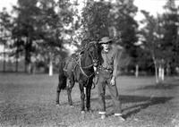 [Unidentified young boy in flat-crowned hat standing with saddled horse, trees in background]