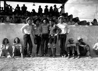 [Five cowboys standing in front of grandstand; two women sitting to left & three men to the right]
