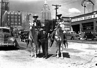 [Bob Wills & Johnnie Lee Wills on horses wearing silver saddles in city street with Mayo standing]