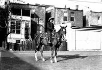 [Unidentified cowgirl on horse with row of houses behind]