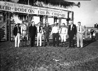 [Group of nine cowboys in various western wear in front of chutes at Billy Crosby's Rodeo]