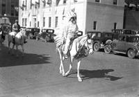[Unidentified Cowgirl atop horse in western clothing and feathered headdress on street in parade]