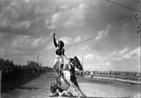 [Unidentified Cowgirl standing on saddle of sitting horse waving with her right hand]