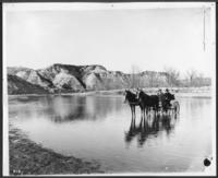 [2 men in a horse drawn buggy crossing a river]