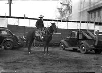 [Unidentified Cowboy holding rope to side atop horse between cars and truck]