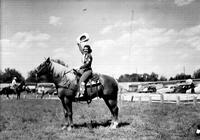[Unidentified cowgirl contestant atop horse raising her hat]