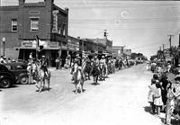 [Two cowgirls with flags lead group of riders down city street in front of "City Drug Co."]