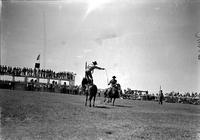 [Unidentified Cowboy atop horse and throwing rope loop in front of approaching horse and rider]