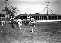 [Majorette leading cowboy baton twirler and marching band in arena]