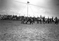 [Indian dancers look on as two Indian men pull pants off rodeo clown in arena]