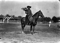 [Col. Jack King in wooly chaps, wearing a large hat and carrying a bouquet on horseback]