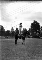 [Unidentified Cowgirl standing on stationary horse spinning a large vertical rope loop to her side]