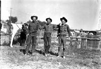 [Three unidentified cowboys (possibly related) standing in front of slat fence]