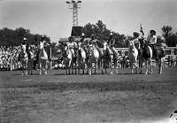 [Cowboys & cowgirls on horseback in line in arena with Chutes behind them]