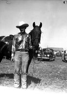 [Unidentified Cowboy standing with horse with notched ear, autos in background]