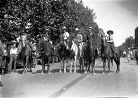 [Five unidentified cowboys and a cowgirl (far right) on horseback in street]