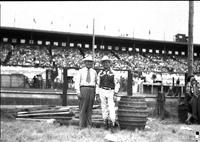 [Autry standing with unidentified man in arena next to clowns' barrel, grandstand in background]