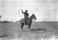 [Unidentified Cowboy holding rope loop aloft in field while atop horse]
