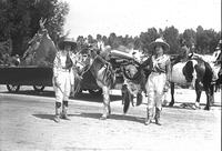 [Two Unidentified Cowgirls prepare for parade standing next to mule, Tipi on float to left]