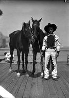 [Possibly Billy Keene in fancy two-toned western outfit holding leads of two unsaddled horses]