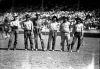 [Six cowboys, three with coiled ropes, standing in arena in front of grandstand]