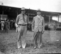 [One man in business suit, hat and with cane standing besides older cowboy with necktie]