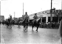 [Unidentified Cowboy and Cowgirl on horseback & side by side leading group of others down street]