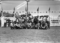 [Group of standing & squatting cowboys in front of chutes; One cowboy on horseback in center]