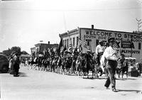 [Large group of cowgirls on horseback & carrying flags parading down street]