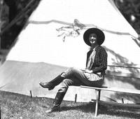 [Unidentified cowgirl sitting on bench by tipi]
