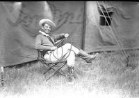 [Unidentified Man sitting in chair by tent holding whip]