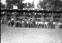 [Group of cowboy contestants standing in front of chutes; rodeo clowns off to the left of group]