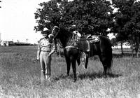 [J. Wills standing by dark horse wearing silver saddle.  Trees and Stadium in background.]