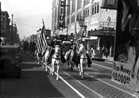 [Pair of mounted cowboys bearing American Flags and leading riders down street in front of stadium]