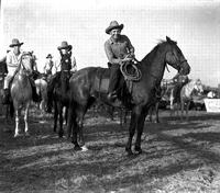 [Laid-back unidentified cowboy on horseback, megaphone in hand; other cowboys on horses behind]