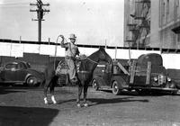 [Unidentified Cowboy with rope loop aloft atop horse between car and truck]