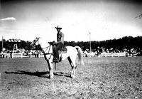 [Unidentified Cowboy on horse in grass area; fence, people, and trees in background]