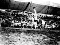 [Unidentified cowboy doing shoulder stand on galloping horse passing canopy-covered stands]