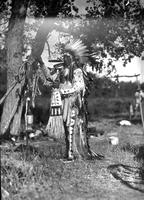 [Indian man in floor length full feathered headdress & costume standing by tree & holding bow]