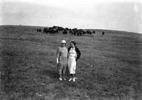 [Two formally dressed unidentified ladies standing in field where Buffalo graze]