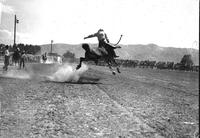 [Unidentified Cowboy riding bronc with Spectators in distant stands, trees, and hills in background]