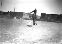 [A hatless Bill Linderman riding and staying with his bronc "Iron Mountain" in near empty arena]