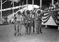 [Three Indian men in full feathered headdress standing in front of grandstand]