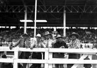 [Group of five unidentified men in western attire behind fence with spectators in background]