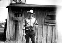 [Unidentified Cowboy standing in front of wooden building holding coil rope in left arm]