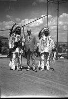 [Man in business attire holding a "Fair-Program" standing between two Indian men in native clothing]