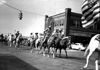 [Unidentified Cowboys, Cowgirls & young riders moving in pairs down city street]