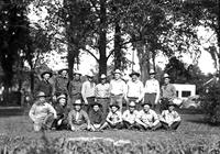 [Group of sitting and standing Cowboys in front of trees]